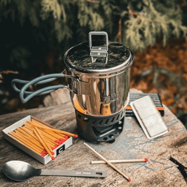 Esbit titanium stove with pot and matches on a wooden surface in a forest setting. Photo by Rigad.