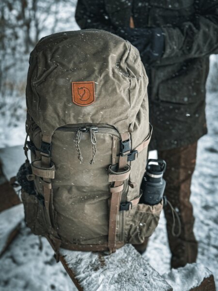 Fjällräven backpack in a winter setting, covered with snow, with a bottle in the side pocket and a figure in gloves standing in the background. Photo: Rigad