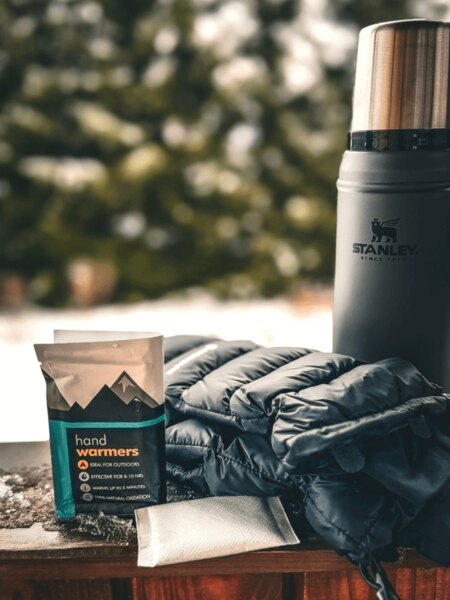 Haago hand warmers, a packed down jacket, and a Stanley thermos placed on a snow-dusted table in the winter nature. Photo: Rigad