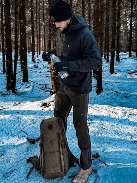 A tourist in the winter forest pours a hot drink from a thermos, with a Fjällräven trekking backpack at their feet. Photo: Rigad