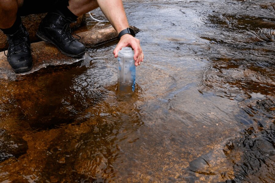 A person collecting water from a stream into a soft bottle with a squeeze filter during an outdoor trip. Photo: Rigad.