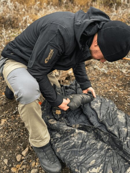 Man in a black jacket packing a camouflage sleeping bag on rocky ground.