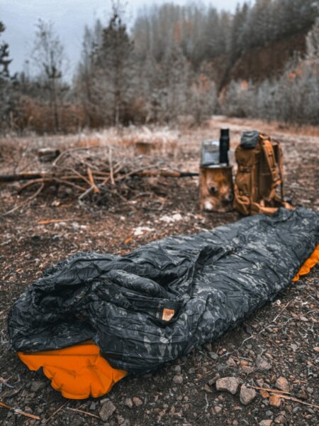 Unzipped camouflage sleeping bag on the ground in an autumn landscape, with a backpack in the background.