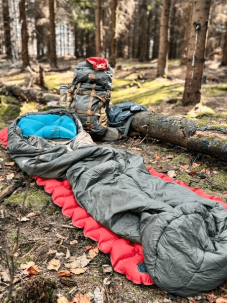 Green-and-gray sleeping bag on a red inflatable sleeping pad, with a blue pillow and backpack in a forest.