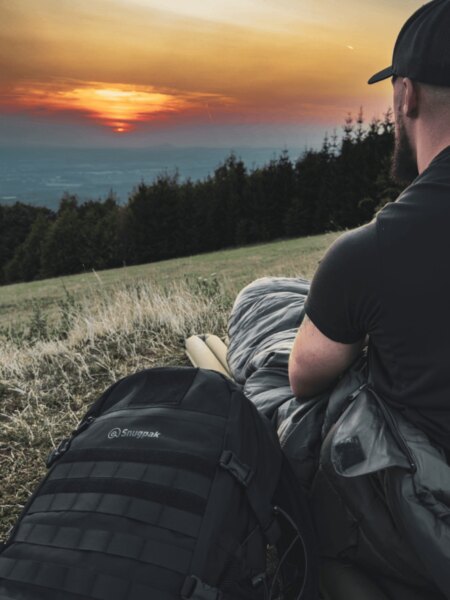 Man sitting in a sleeping bag on a grassy hillside at sunset, with a black backpack beside him.