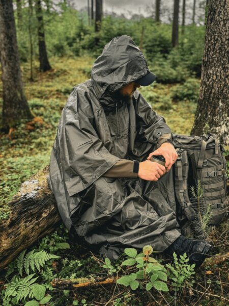 Man wearing a Helikon-Tex waterproof poncho while sitting beside a tactical backpack in wet woodland.