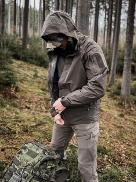 Man wearing a hooded Helikon-Tex Chinook waterproof jacket beside a camouflage backpack in woodland.