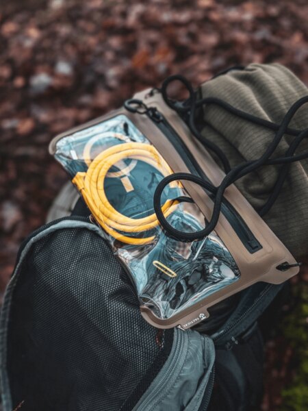 Transparent Tactical water-resistant pouch containing electronic devices and a yellow charging cable on an open backpack.