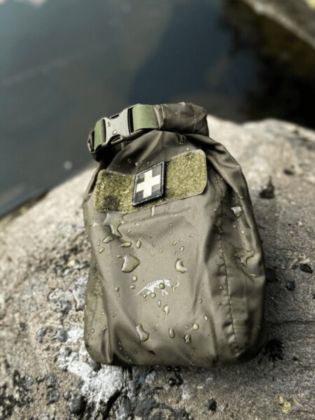Olive-drab Tasmanian Tiger roll-top dry bag with a medical identifier on a wet rock beside the water.