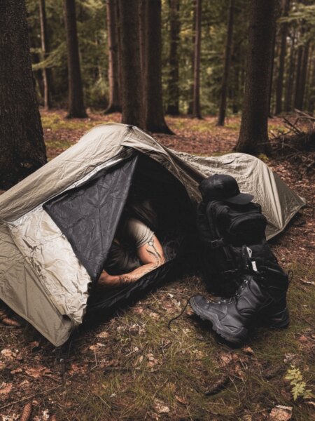 Person lying inside a low-profile bivy shelter with the entrance open in a forest, with a backpack and boots beside it.