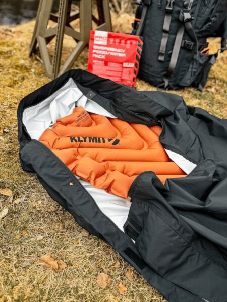 Orange Klymit inflatable sleeping pad inside an open black bivy bag, with a backpack and cooking equipment in the background.