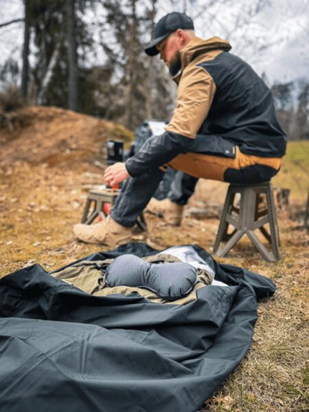 Man sitting on a folding stool in a forest, with a complete sleep system, protective bivy bag, and pillow laid out in the foreground.