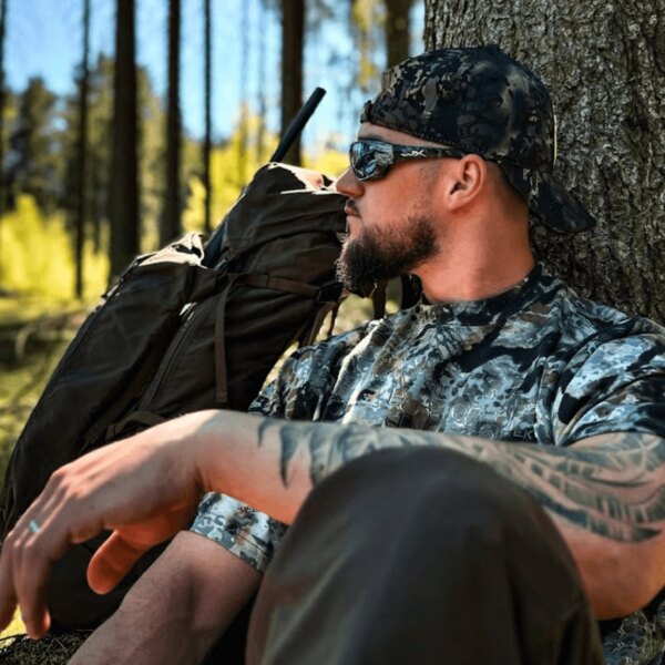 Man wearing Kryptek camouflage gear, seated by a tree with a backpack in a natural outdoor setting. Photo by Rigad.