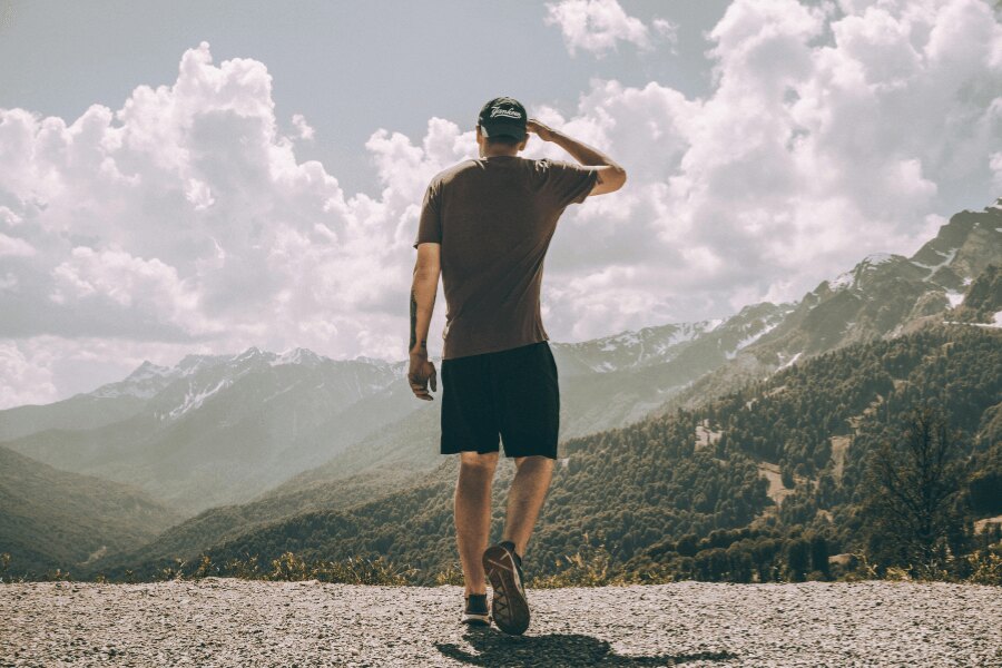 Man in a T-shirt and shorts standing with his back to the camera, overlooking a mountain landscape. Photo: https://www.pexels.com/photo/man-mountains-nature-sky-2577706/