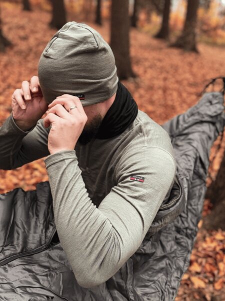 Person in a merino hat and gray thermal shirt sits in a sleeping bag in the autumn forest; the quiet first layer reduces thermal and scent traces. Photo: Rigad