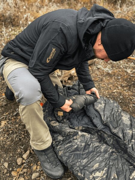 A person in a matte jacket compresses a sleeping bag at the bivouac on the rocky ground; subdued colors and neat gear attachment without rattling. Photo: Rigad