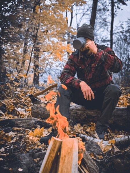 The man in the checkered jacket sits by the fire in the autumn forest and drinks from a metal mug. Autumn camping with the scent of wood and the peace of nature. Photo: Rigad