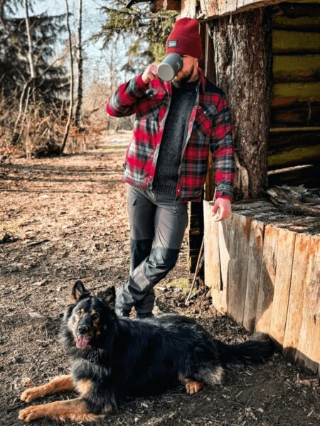 A man in a red jacket and cap is drinking from a mug at a wooden cabin, with a dog lying at his feet. Autumn camping with natural comfort and a loyal companion. Photo: Rigad