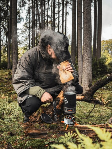 A man in an insulated jacket is kneeling in the forest by a Jetboil stove, from which steam is rising. He is preparing a warm meal or drink as a source of energy and warmth during an autumn expedition. Photo: Rigad