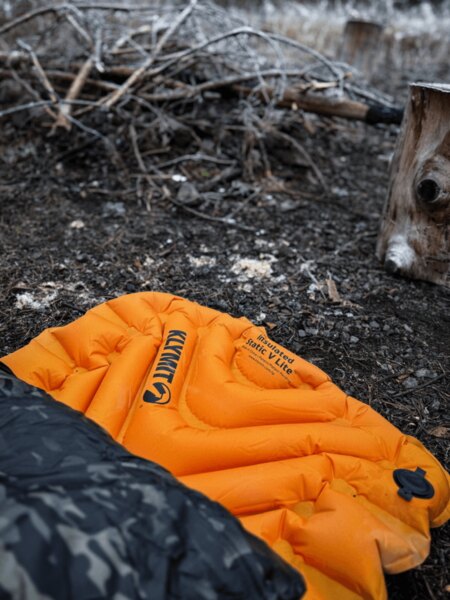 Inflatable sleeping pad Klymit Insulated Static V Lite in orange color on the ground by the campfire, partially covered by a sleeping bag with a camouflage pattern. Insulation from cold and moisture when sleeping outdoors. Photo: Rigad
