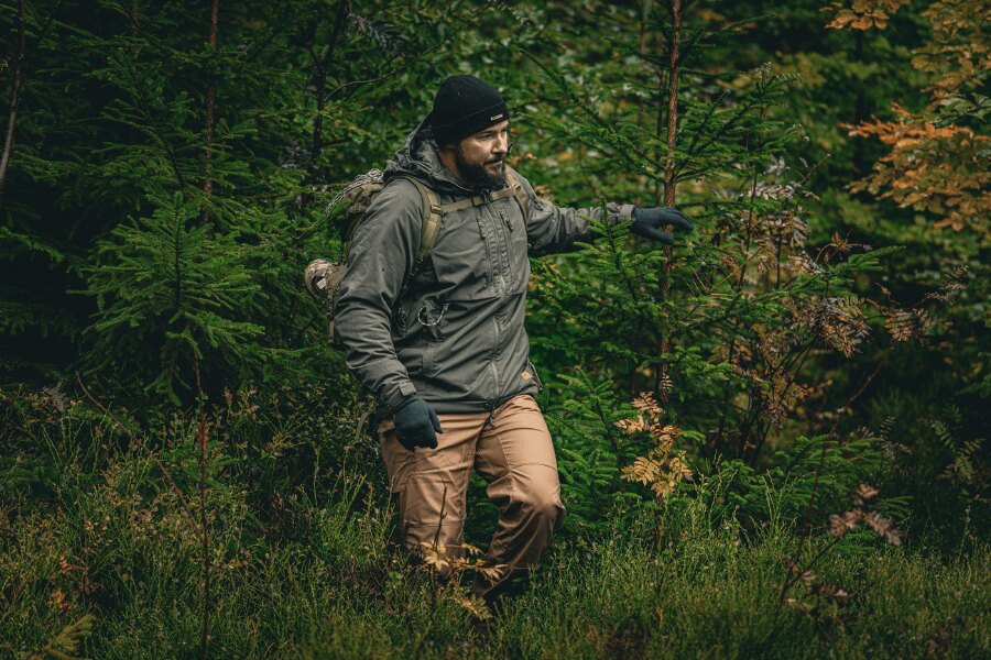 Man in an autumn forest in a gray winter jacket Yukon with Climashield™ Apex<sup>&reg;</sup> insulation from Helikon-Tex<sup>&reg;</sup>, wearing a cap and gloves, with a backpack; demonstration of proper layering and protective outer layer for wet and windy conditions. Photo: Rigad