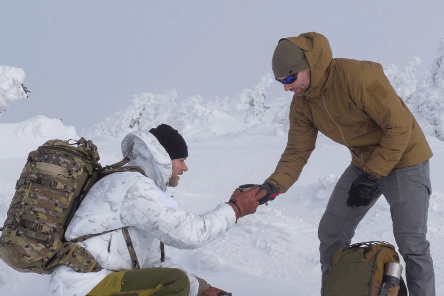Two men in a winter landscape during a break on the snow – one handing the other a hot drink as part of hypothermia prevention. Photo: Rigad