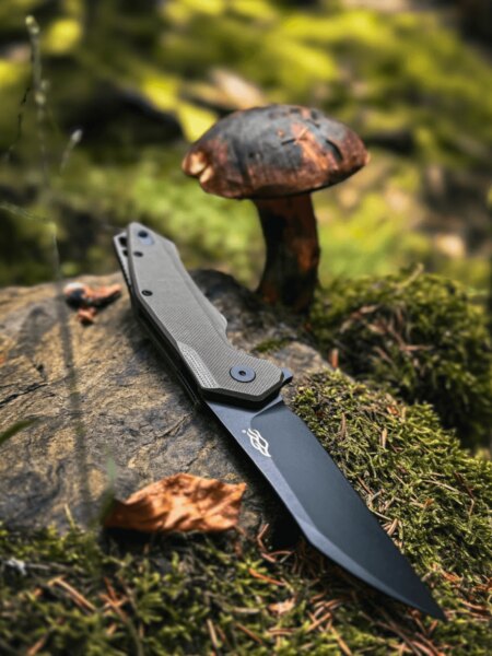 Ganzo folding knife resting on a rock in the forest beside a mushroom.