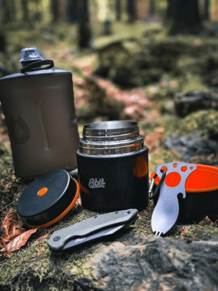 Open Esbit insulated food jar with an outdoor bottle, folding knife and cutlery in a forest setting.
