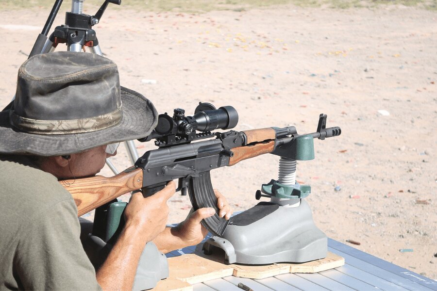 Shooter aiming a Romanian WASR-series rifle fitted with a riflescope and muzzle brake at an outdoor range.
