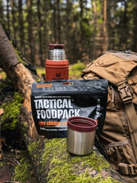 A Tactical Foodpack freeze-dried meal with a Stanley thermos, cup and backpack in a forest.