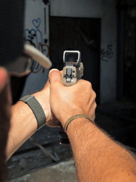 Shooter aiming a pistol equipped with a red dot sight, viewed over the shoulder through the optic window with the red dot visible.
