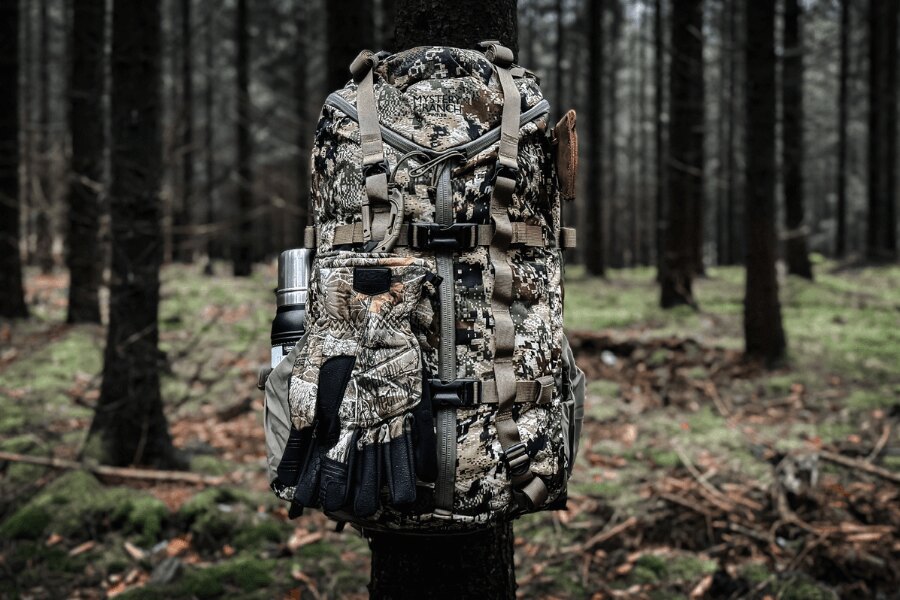 Mystery Ranch 2 Day Assault backpack in a camouflage pattern with attached gloves and a water bottle in woodland.