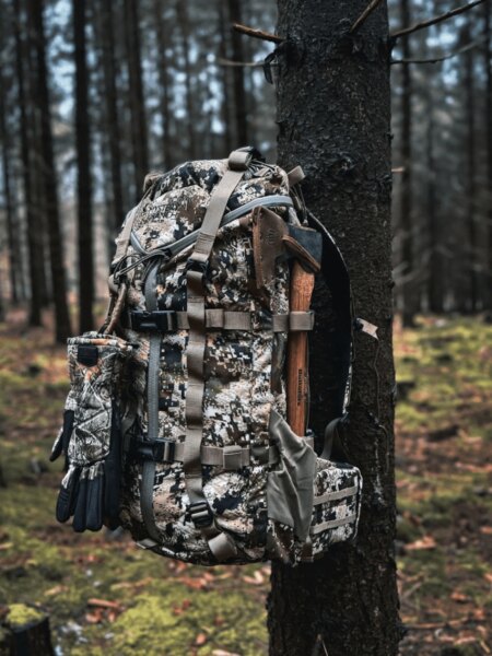 Mystery Ranch 2 Day Assault backpack in a camouflage pattern with an attached axe, gloves, and pouches beside a tree.