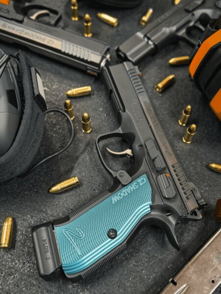 CZ Shadow 2 competition pistol with blue aluminum grip panels displayed among ammunition and shooting gear.