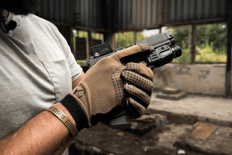 Shooter wearing Mud Brown Dýlejc Bivoj gloves and holding a pistol fitted with a red-dot sight and weapon light in a two-handed grip.
