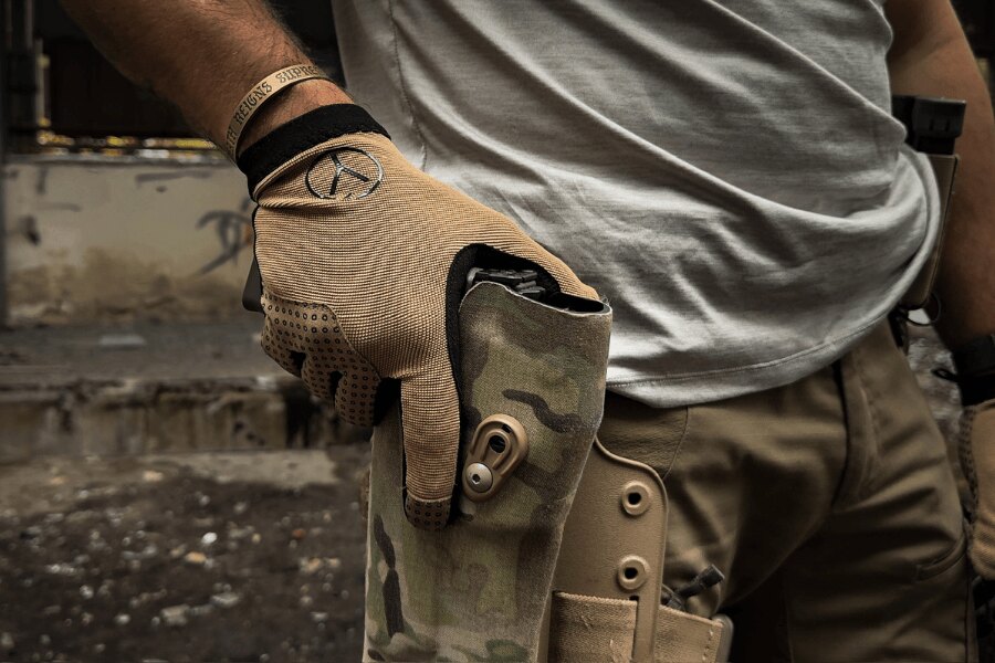 Hand wearing a Mud Brown Dýlejc Bivoj glove resting on a pistol in a belt-mounted holster.