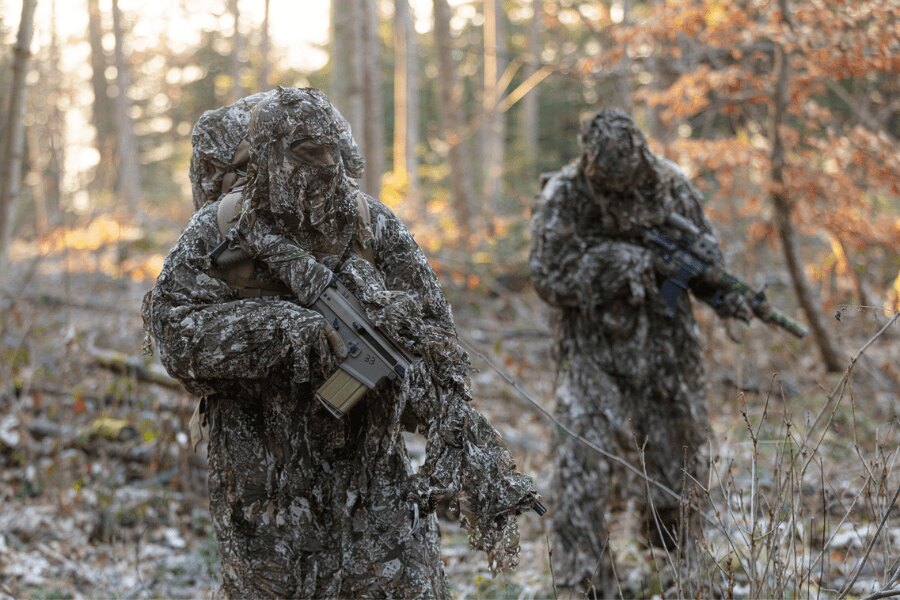 Two armed users wearing GHOSTHOOD camouflage overgarments in an autumn forest.