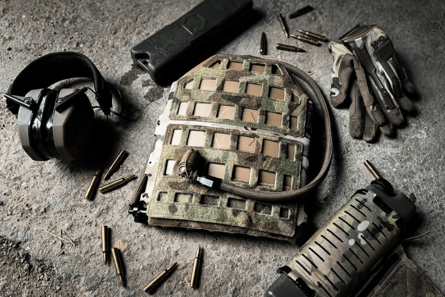 Qore Performance ICEPLATE hydration system in a camouflage cover with a drinking tube, positioned beside gloves, hearing protection, and other equipment.