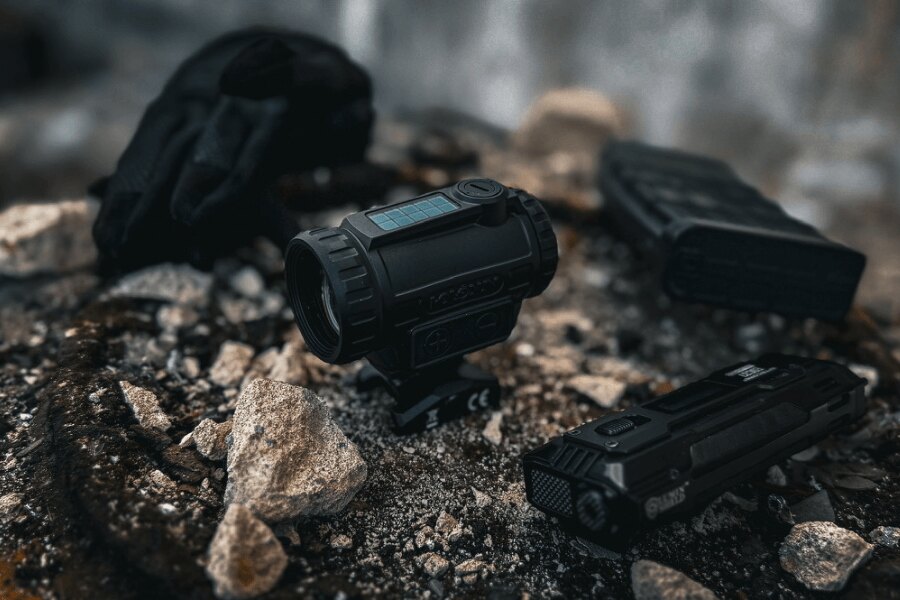 Holosun tube-style red dot sight and IRIS laser module placed on rocky ground beside gloves and a magazine.