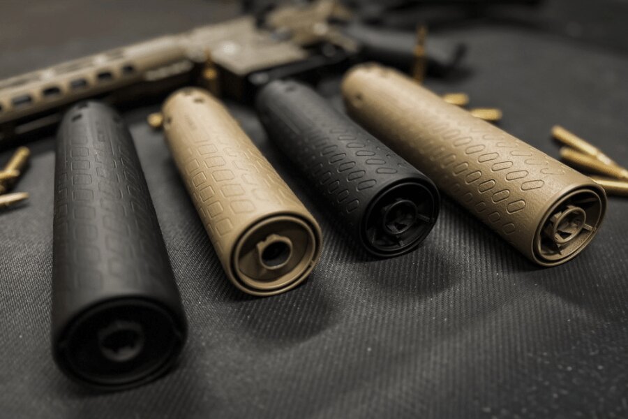 Four black and tan firearm suppressors arranged on a dark mat, with a rifle and ammunition in the background.