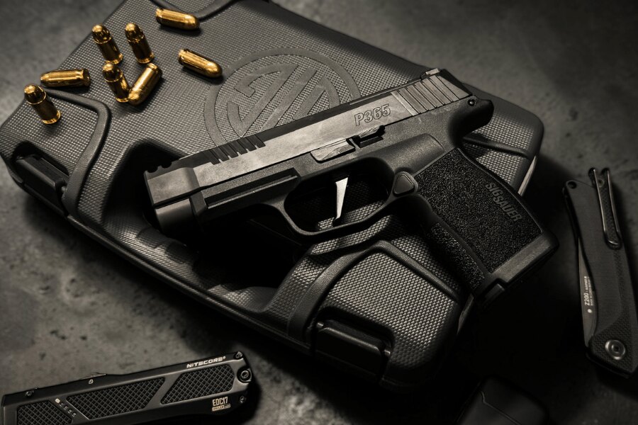 Black SIG Sauer P365 pistol displayed on a branded hard case among cartridges, a folding knife, and a flashlight.