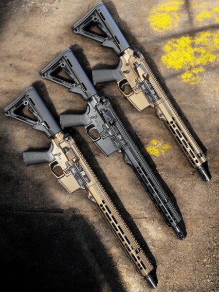 Close-up of a black AR-15 with its ejection-port cover open, positioned between two tan rifles.