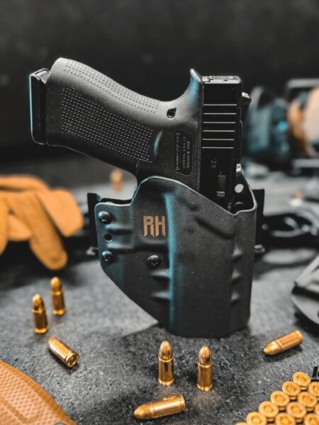 A Glock pistol in a black RH Holsters OWB holster, standing on a table among ammunition and shooting gear.